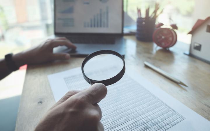 A magnifying glass looking over a spreadsheet, with an open laptop in the background.